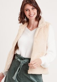 Gilet en fausse fourrure beige avec une texture douce, doté d'une fermeture zippée et d'un col. Associé à un pull blanc et à une jupe en velours côtelé verte.