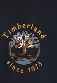Tummansininen puuvillapaita, jossa on graafinen kuvasto tyylitellystä puusta, ja kultaiset tekstit "Timberland" ja "since 1973".