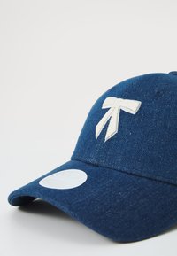 Ναυτικό μπλε καπέλο από τζιν με μια κεντητή λευκή κορδέλα μπροστά και μια κυκλική ετικέτα λογότυπου στη γείσο.