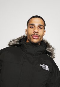 Zwarte geïsoleerde parka-jack met een met bont gevoerde capuchon, voorzien van een gladde textuur en een logo op de linkerkant van de borst. Ritssluiting, ontwerp met hoge kraag.