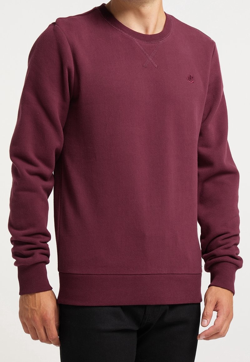 Homme portant un sweat-shirt bordeaux à manches longues avec col rond, poignets et ourlet côtelés, associé à un pantalon noir.