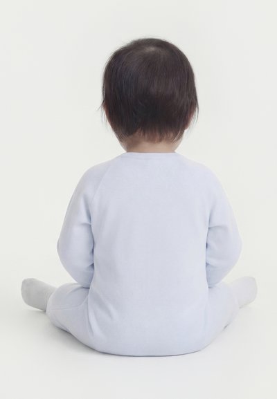 Νήπιο με σκούρα μαλλιά που φοράει ανοιχτό μπλε πυτζάμες, καθισμένο στο πάτωμα με την πλάτη γυρισμένη σε λευκό απλό φόντο.