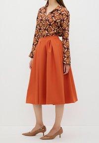 Långärmad blus med ett flerfärgat abstrakt mönster, kombinerad med en knälång orange plisserad kjol och nudefärgade lackskor.