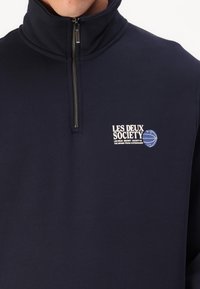 Sudadera de forro polar azul marino con un pequeño logo blanco en el lado izquierdo del pecho y una textura suave. Cuenta con una cremallera de metal y un cuello alzado.