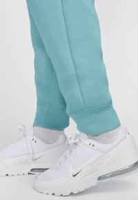 Svijetloplave trenirke s manžetama na gležnjevima, uparene s bijelim Nike tenisicama koje imaju mrežaste i kožne teksture, crne naglaske i vidljivo zračno jastučenje.