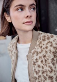 Jeune femme aux cheveux bruns courts portant une veste beige à imprimé léopard texturée, regardant sur le côté sur un fond sombre.