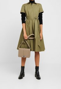 Oliwkowa sukienka koszulowa z krótkimi rękawami i paskiem w talii, w połączeniu z beżową torebką o wyraźnej strukturze i czarnymi botkami za kostkę.