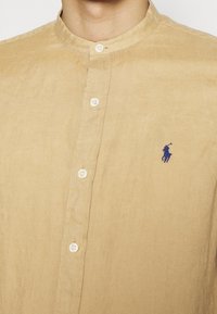 Hnědá košile na knoflíky s límečkem, s bílými knoflíky a malým modrým logem na levé straně hrudi. Měkká, texturovaná tkanina.