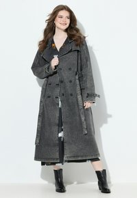 Usměvavá žena s dlouhými vlnitými vlasy, která nosí dlouhý tmavě šedý dvouřadý kabát, černé kozačky a černé vzorované šaty, stojící před bílou zdí.