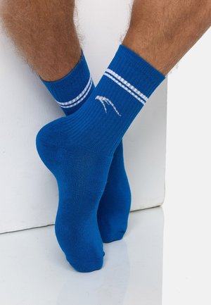 Füße, die hellblaue gerippte Socken mit zwei weißen Streifen und einem kleinen weißen Logo tragen, stehen auf einem glänzenden weißen Boden vor einer weißen Wand.