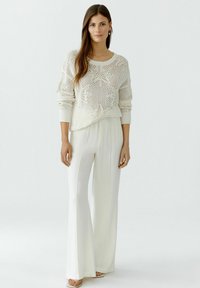 Cremefarbener Strickpullover mit lockerer Passform und Sternenmuster, kombiniert mit hochtaillierten, weit geschnittenen weißen Hosen und flachen Sandalen.