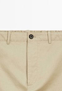 Pantalons en coton beige avec une fermeture à bouton noir, un devant plat et des passants de ceinture. Texture lisse avec des détails de construction sur mesure.