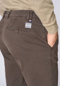 Pantaloni in cotone marrone con tasca posteriore e chiusura a bottone. Presenta un'etichetta con logo, dettagli cuciti e una superficie liscia.