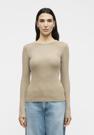 Γυναίκα με μακριά σκούρα μαλλιά φορώντας ένα μπεζ ριμπ πλεκτό μακρυμάνικο τοπ και ανοιχτό μπλε τζιν, στέκεται μπροστά σε απλό λευκό φόντο.