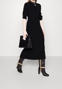 Schwarzes geripptes Strickkleid mit Ellbogenärmeln, kombiniert mit knielangen schwarzen Lederstiefeln mit beigen Akzenten und einer schwarzen Tasche mit einem runden Griff.