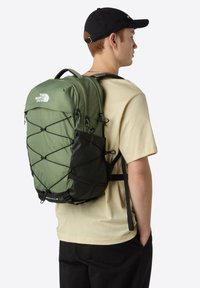 Green nylon backpack with black accents, featuring multiple compartments, mesh side pocket, and bungee cord design for added storage.