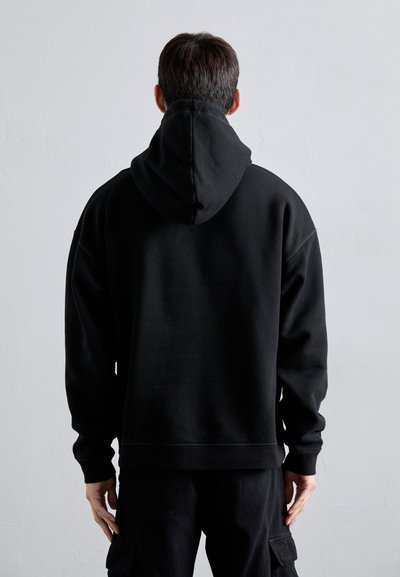 Schwarzer Hoodie mit einer großen, gefütterten Kapuze. Lockerer Schnitt, gerippte Bündchen und Saum. Glattes, weiches Material. Getragen mit passenden dunklen Hosen.