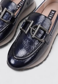 Los mocasines de charol navy presentan una prominente hebilla de metal, una puntera redondeada y una suela gruesa con un acabado texturizado.