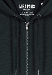 Felpa in cotone verde scuro con zip frontale, dotata di cappuccio regolabile con coulisse e dettagli metallici argentati, etichettata con marchio e istruzioni per la cura.