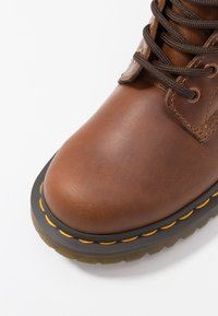 Botas de cuero marrón con punta redondeada, cordones marrones, detalles de costura naranja y una suela gruesa de goma negra con un patrón texturizado.