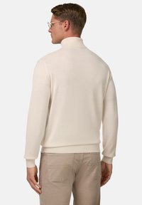 Krēmkrāsas uzvalks ar augstu kakla izgriezumu, izgatavots no mīksta adījuma materiāla, ar pieguļošu siluetu un ribotām piedurknēm, saskaņots ar gaiši bēšām biksēm.