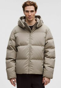 Junger Mann mit lockigem Haar, der eine beige Steppjacke mit Kapuze trägt und vor einem schlichten hellen Hintergrund steht.