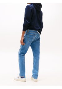 Personne portant un jean bleu clair, des baskets blanches et un sweat à capuche bleu marine, debout de dos sur un fond blanc uni.
