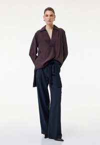 Mujer con una blusa suelta de color púrpura oscuro con escote en V profundo y pantalones anchos azul marino atados a la cintura, posando frente a un fondo blanco.