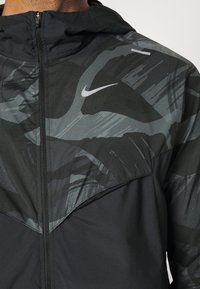 Nike hoodie met een zwart en grijs camouflagepatroon, lichtgewicht stof, textuur aan de bovenkant en een ritssluiting aan de voorkant.