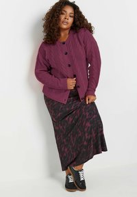 Cardigan en maille torsadée violette avec des boutons noirs, porté sur une longue jupe sombre ornée d'un imprimé léopard rose et noir, associé à des baskets noires.