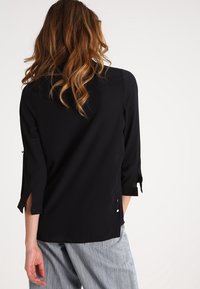 Blusa nera con maniche a tre quarti, texture morbida, polsini doppi con bottoni e spacco sui lati per facilitare i movimenti. Vista posteriore.