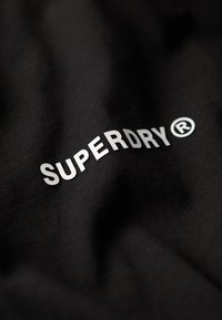 Zwarte stof met een zachte textuur, waarin het zilveren logo "SUPERDRY ®" prominent is weergegeven in een gestileerd lettertype.