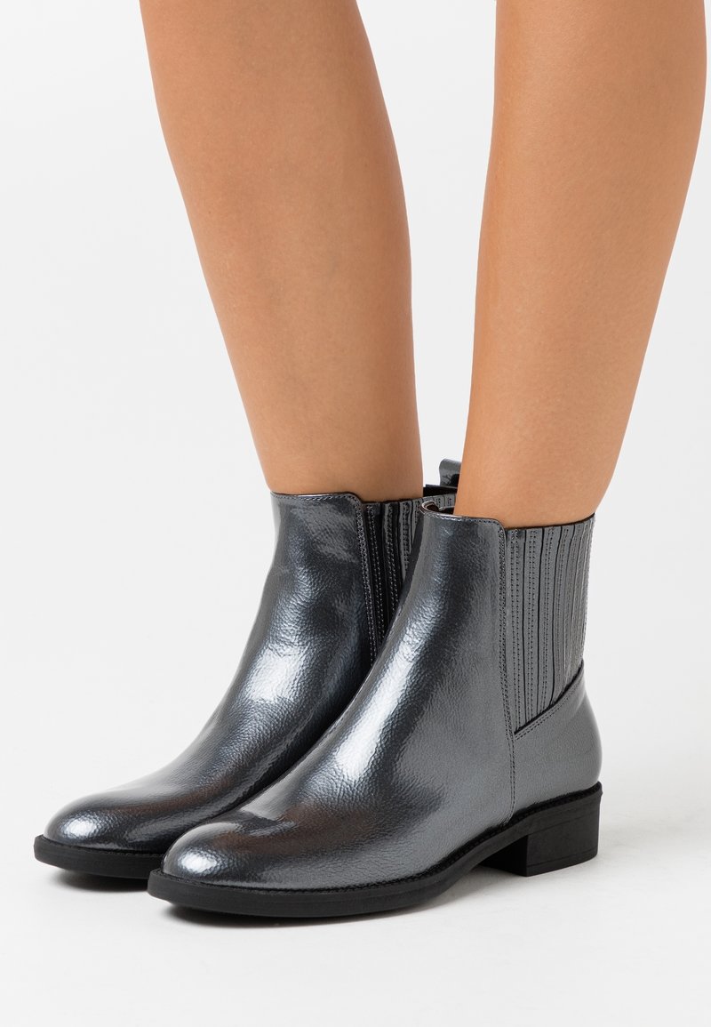 Czarne skórzane buty do kostek z błyszczącym wykończeniem i bocznymi zamkami noszone na gołych nogach, stojące na białej powierzchni.