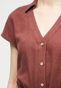 Chemise rouille à col, à manches courtes, avec un col en V et trois boutons ronds en bois visibles sur le devant.