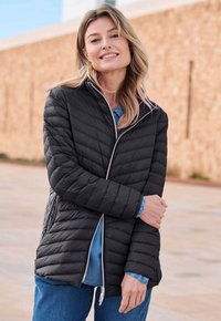 Lachende vrouw in een zwart gewatteerd jack over eendenim blouse en blauwe jeans, staand buitenshuis tegen een gestructureerde beige muur.