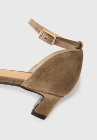 Sandale à talon en suède marron avec une bride de cheville, une boucle en argent et un petit talon carré. Intérieur lisse et design incurvé à l'arrière.