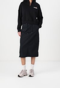 Musta vetoketjullinen collegepaita, jossa pieni logo rinnassa, yhdistetty mustaan taskurullahameeseen. Malli käyttää vaaleanpunaisia tennareita ja valkoisia sukkia.