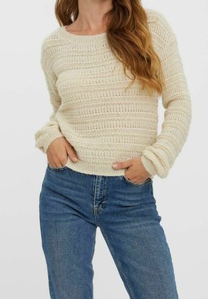 Kobieta ubrana w kremowy sweter dziergany z fakturą oraz niebieskie jeansy z wysokim stanem, ręce schowane w przednich kieszeniach.