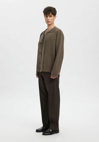 Hnědá pletená košile s límečkem, předními knoflíky a dvěma našitými kapsami, doplněná tmavě hnědými kalhotami na míru a černými botami.