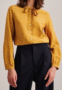Mustard yellow blouse with white leaf patterns, tie-neck detail, long sleeves, and button closure, paired with navy trousers.