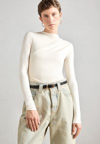 Top blanco de manga larga con un diseño drapeado, combinado con unos jeans de tiro alto de color azul claro y un cinturón negro con una hebilla dorada redonda.
