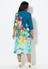 Vestido floral en tonos verde azulado con patrones vibrantes de múltiples colores, mangas tres cuartos y una silueta suelta y fluida; combinado con tacones de color crema.
