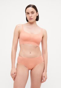 Conjunto de lingerie em tom pêssego, feito de tecido suave, apresentando um design em forma de onda e um padrão texturizado subtil, estilo bralette e cuecas.