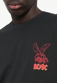 Schwarzes Baumwoll-T-Shirt mit roher gesticktem Motiv einer Fliege und dem "AC/DC"-Logo in stilisierter gelber und roter Schrift auf der linken Brust.