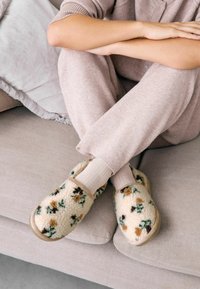 Purūs grietinėlės spalvos šlepetės su žydinčių augalų raštais žalia ir ruda spalvomis, turinčios minkštą viršutinę dalį ir smėlio spalvos guminius padu, dėvimi su šviesios spalvos kelnėmis.