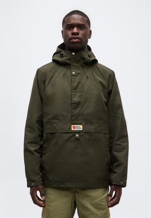 Man met een olijfgroene gewatteerde jas met capuchon en bijpassende broek, staand tegen een effen lichtgrijze achtergrond.