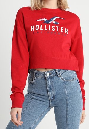 Osoba ubrana w czerwony krótki sweter Hollister California i jasnoniebieskie jeansy z wysokim stanem, stojąca na jednolitym tle.