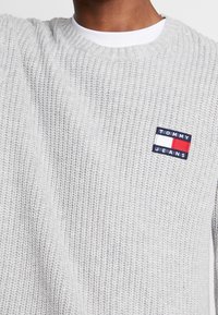 Hellgrauer Strickpullover mit Rundhalsausschnitt, ausgestattet mit einem blauen, roten und weißen Tommy Jeans Logo-Patch auf der linken Seite. Strukturiertes Gewebe.