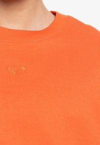 Sudadera naranja con cuello redondo acanalado y un pequeño logo de corazón bordado en el centro, fabricada con material de algodón suave.