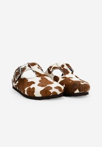 Zapatos de estampado de vaca marrón y blanco con punta redondeada, lados abiertos y una hebilla plateada ajustable. Textura suave y suela negra plana.